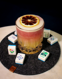 Pink cocktail in a crystal glass with a foamy top and a dried lemon wheel, surrounded by domino tiles on a dark surface.