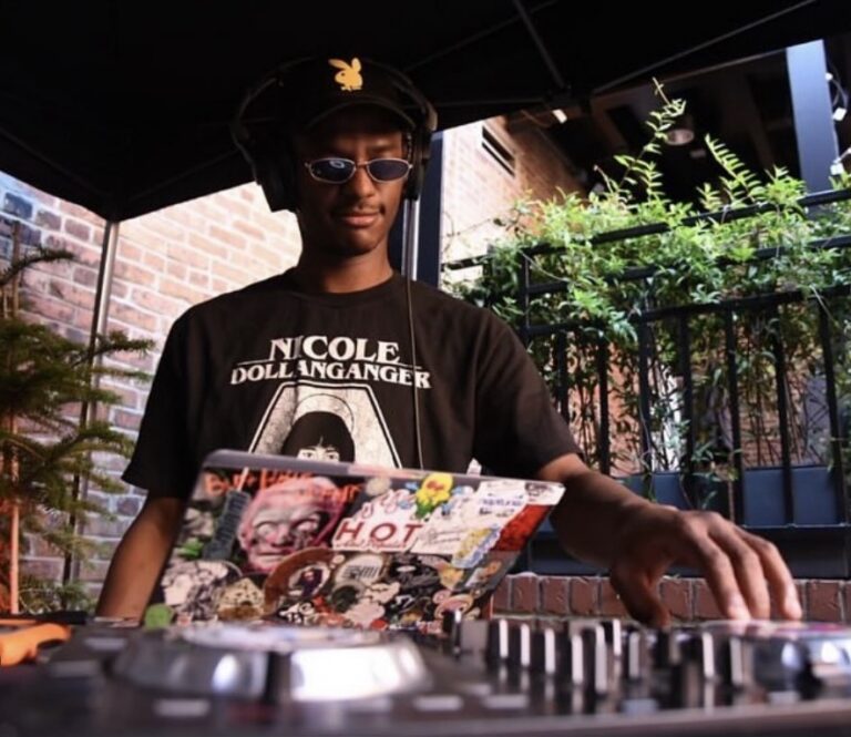 DJ wearing headphones and sunglasses, working at a mixing console with a sticker-covered laptop outdoors under a canopy.