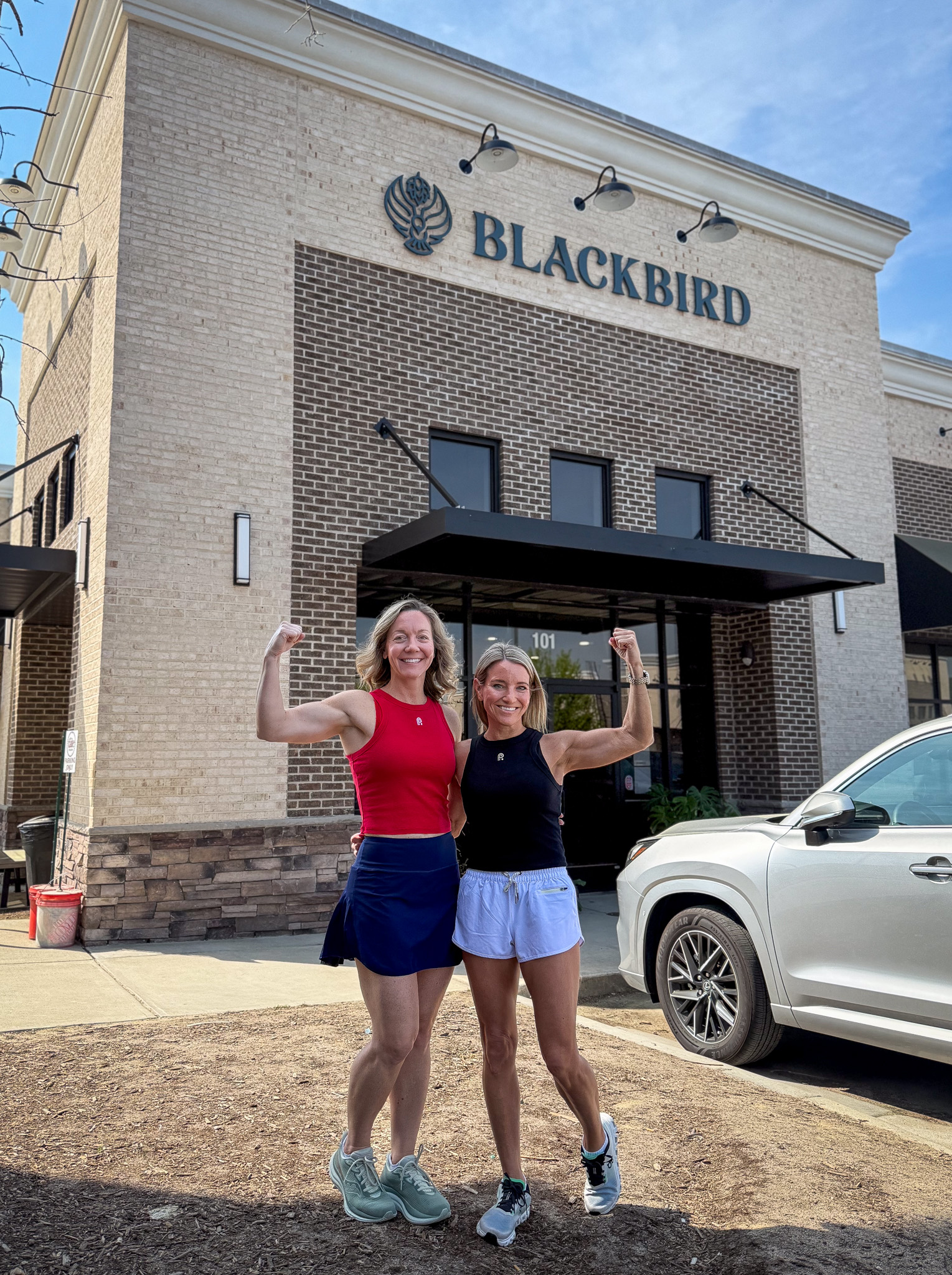 Two smiling women in workout outfits pose with arms raised outside a brick building labeled BLACKBIRD, with a white SUV to the side.