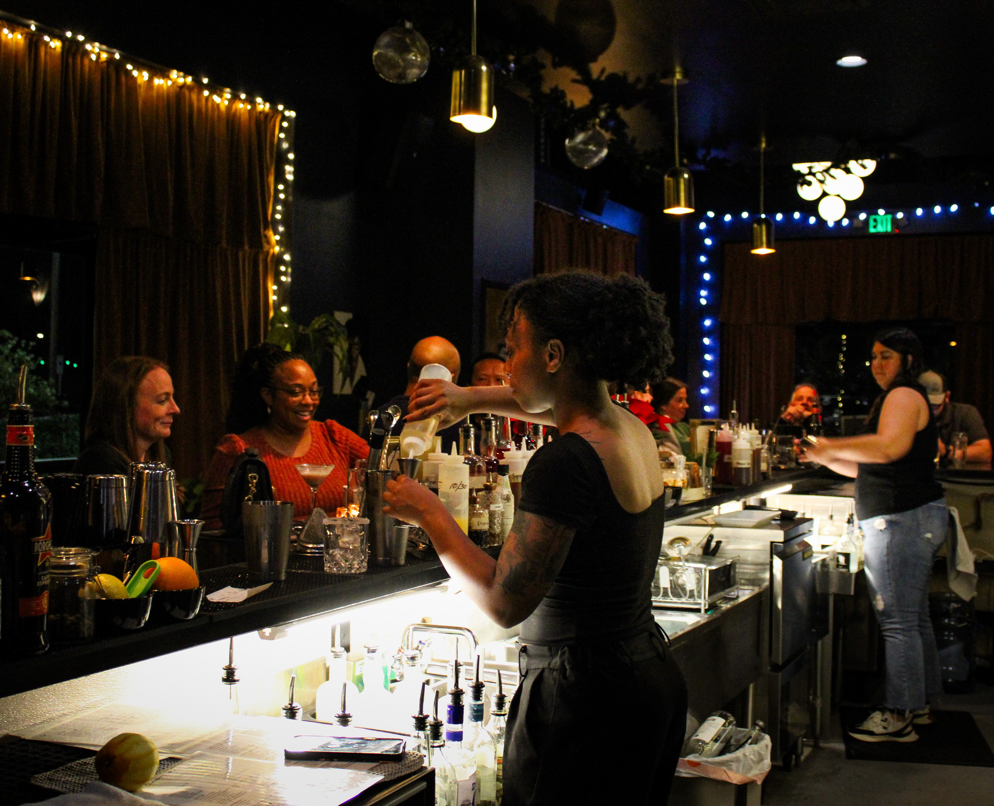 Bartender pours a drink at a dimly lit bar while customers sit and chat, bottles and bar tools visible on the counter.