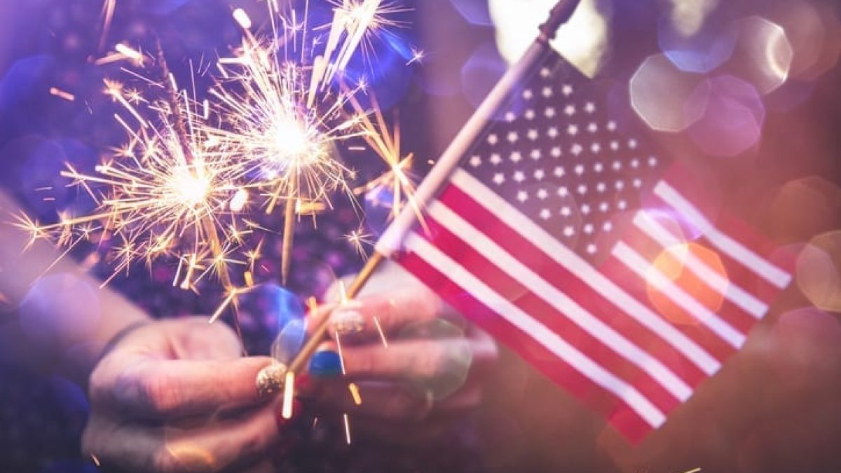 Close-up of a lit sparkler held by hands with a small American flag in the background, conveying celebration and patriotism.