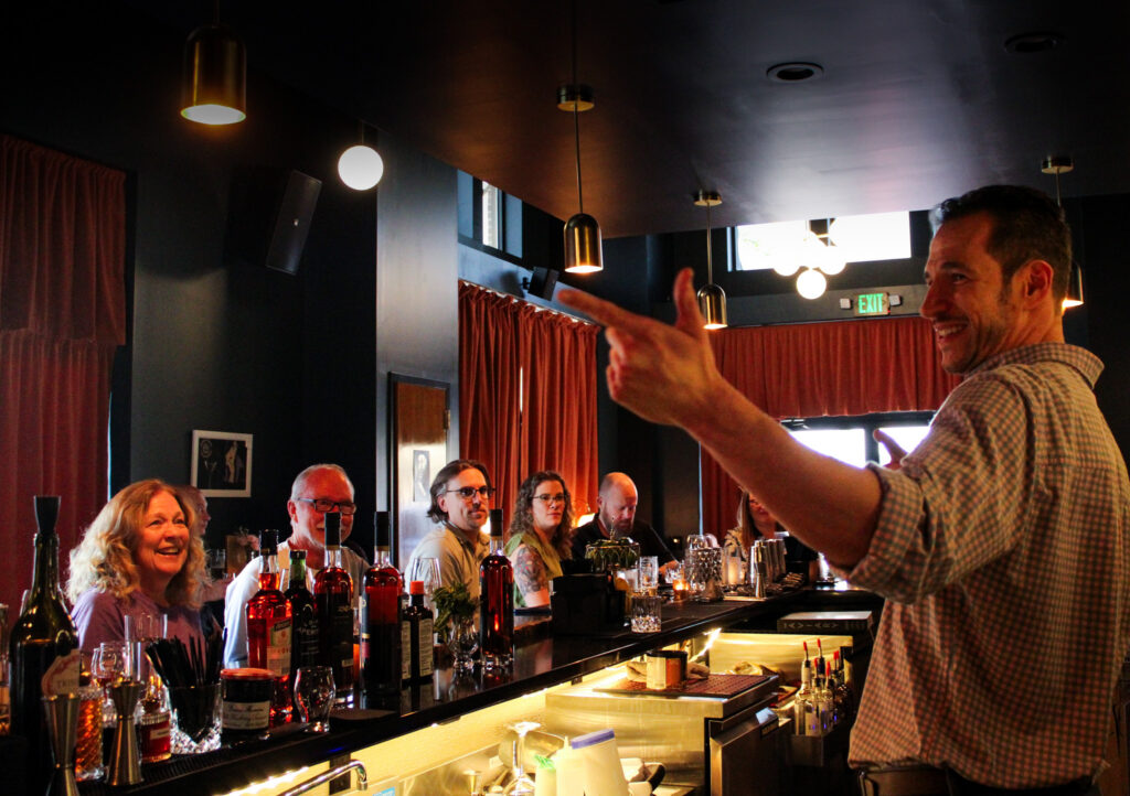 Bartender in a checkered shirt gestures to a group seated at a dark bar with bottles and glasses, warm lighting overhead.