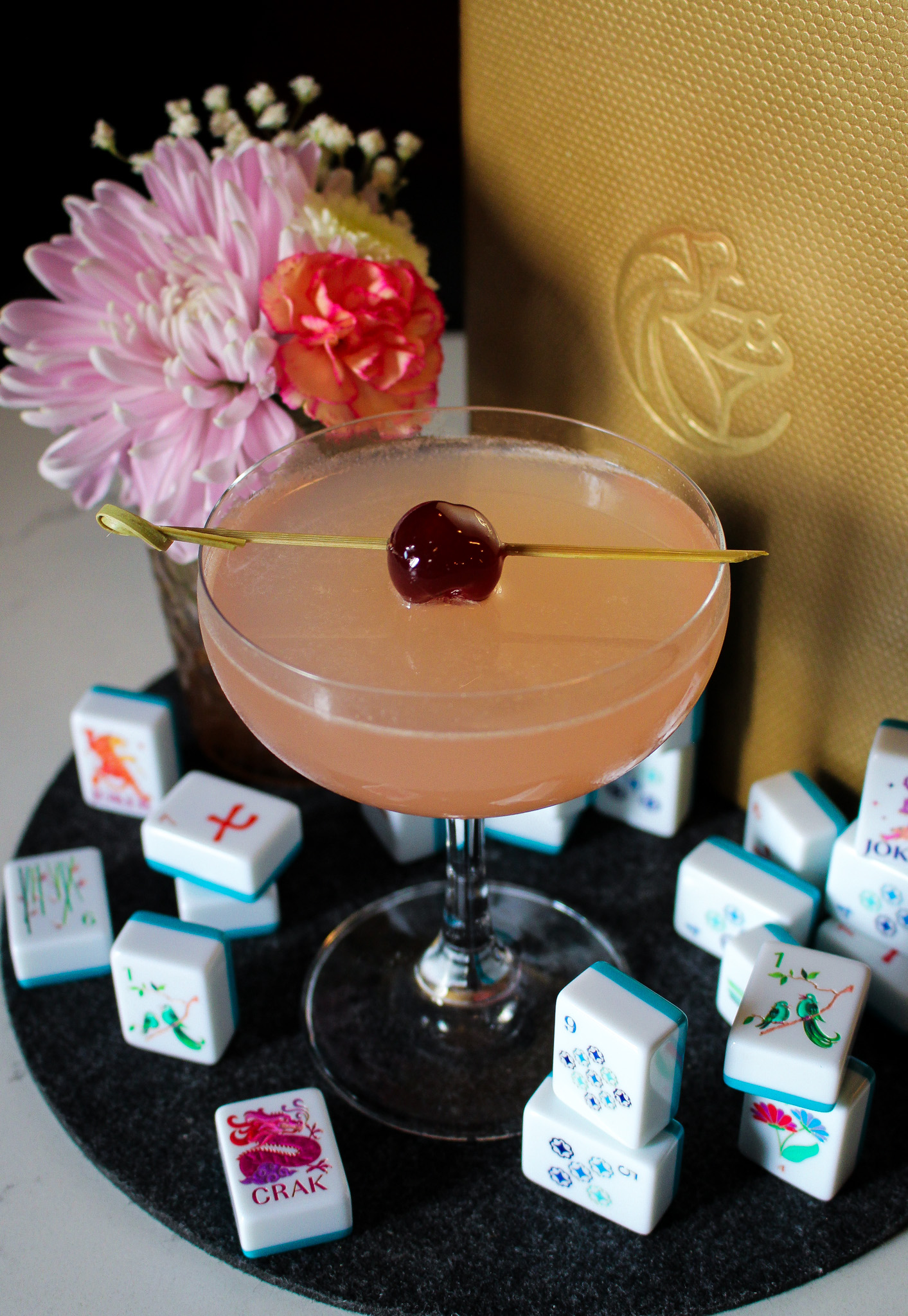 Pink cocktail in a coupe glass with a cherry on a skewer across the rim; blurred pink flowers and a gold textured background, with colorful dice around.